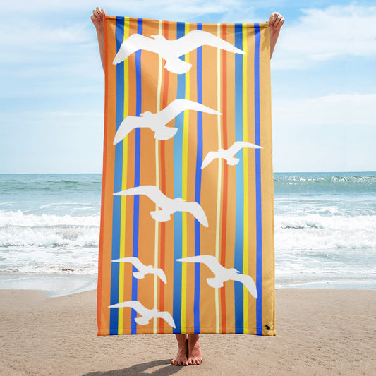 A 30 x 60" Petals & Paws Drift Beach Towel is held up vertically in front of a woman on a sandy beach. A large body of water is in the background. This towel features seagull design with orange striped pattern.