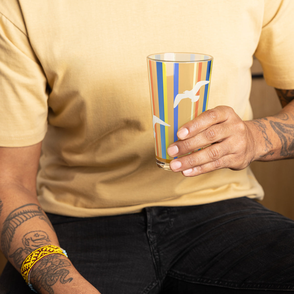 A man holding the Petals & Paws Drift Pint Glass, featuring seagull design with orange striped pattern. Chest and tattooed arms are within the picture.