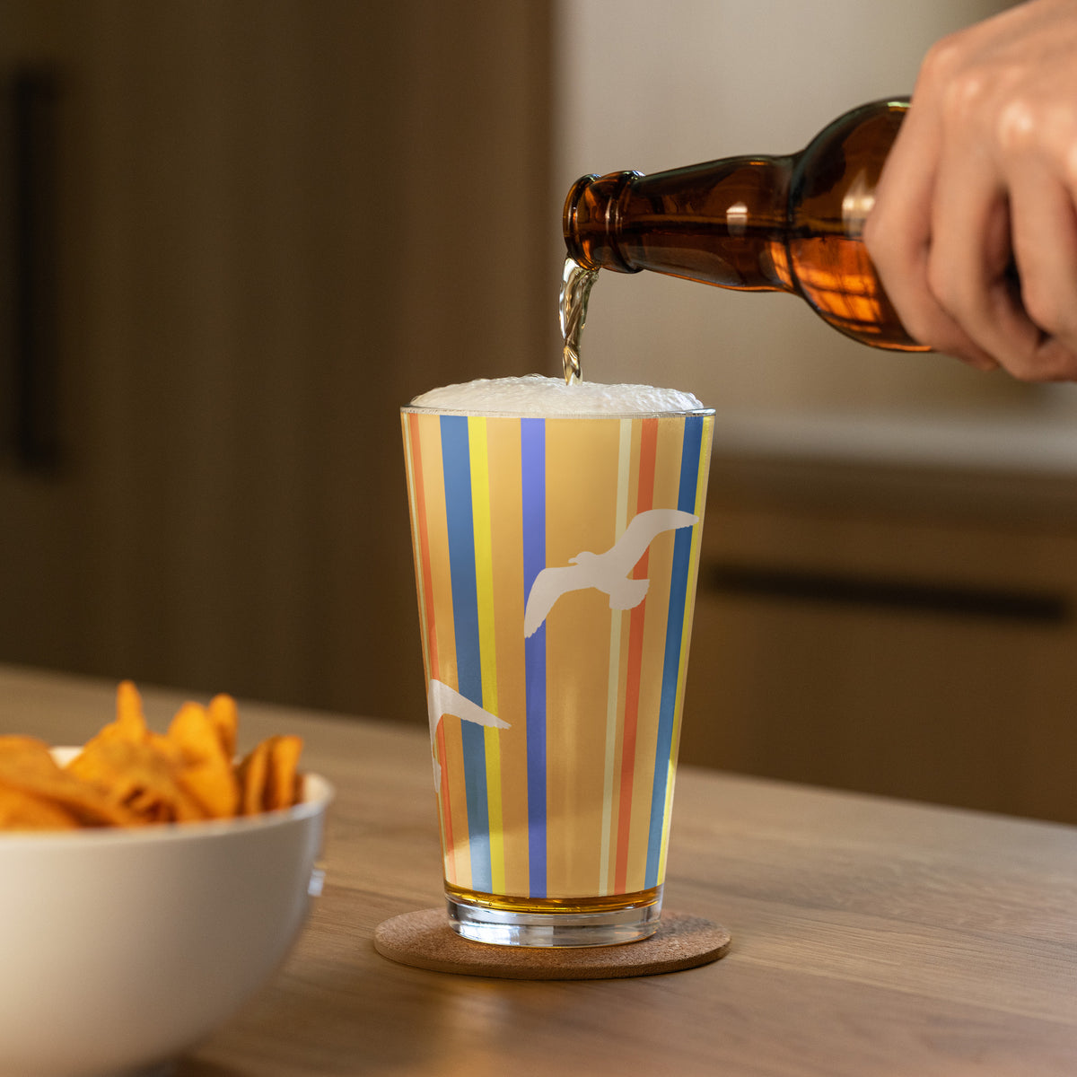 In-action mockup of Petals & Paws Drift Pint Glass, featuring seagull design with orange striped pattern. A hand is pouring beer into the pint glass and a bowl of nachos sits nearby.