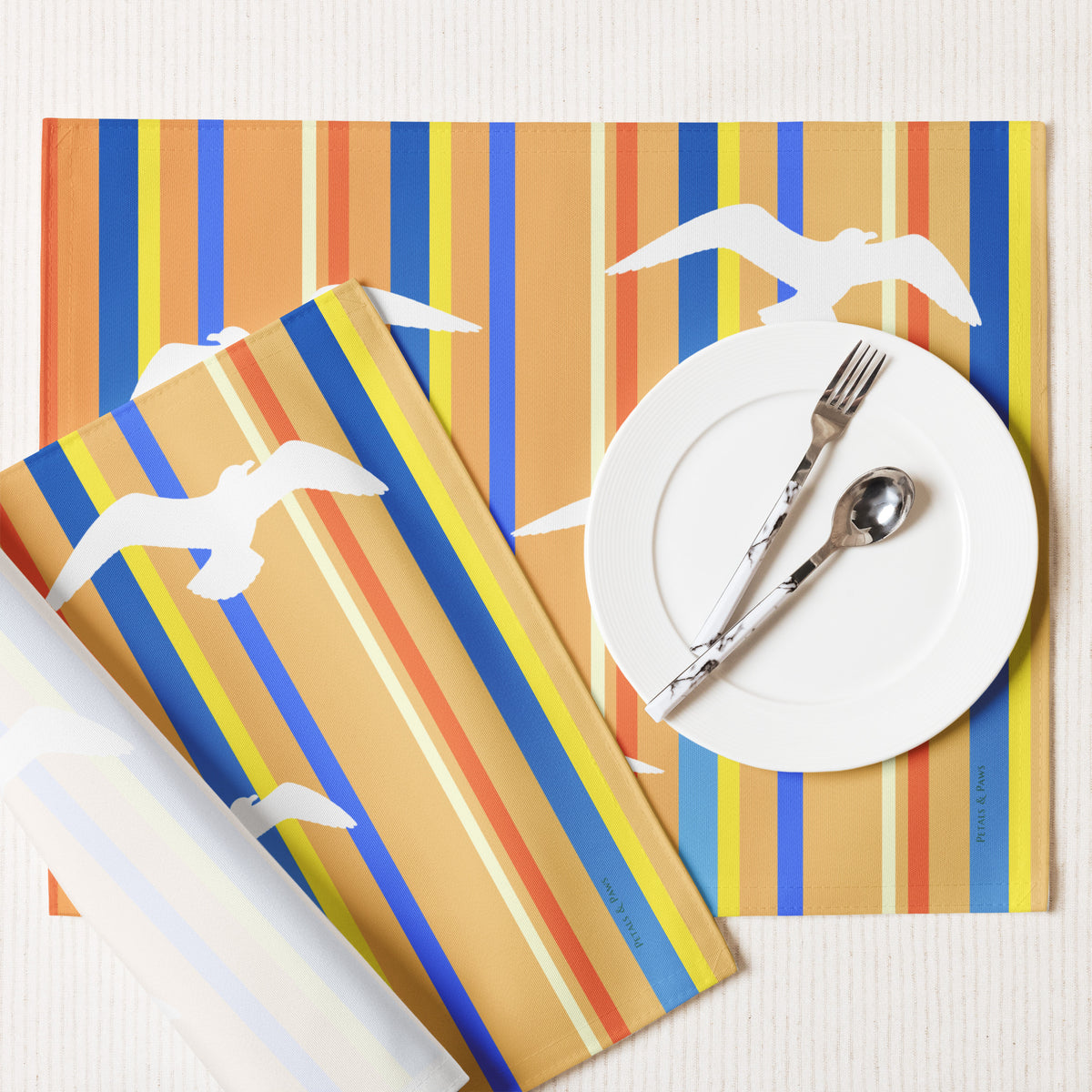 Product mockup of a Petals & Paws Drift Placemat, featuring seagull design with orange striped pattern. This image shows the white back and hemmed edges.