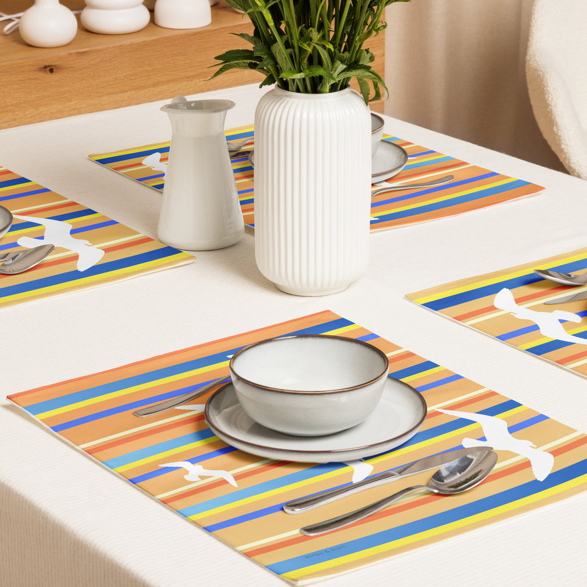 Product mockup of a Petals & Paws Drift Placemat, featuring seagull design with orange striped pattern. This image shows cross-view of a dining table set for four with plates, bowls, utensils, and a vase holding cut flowers.