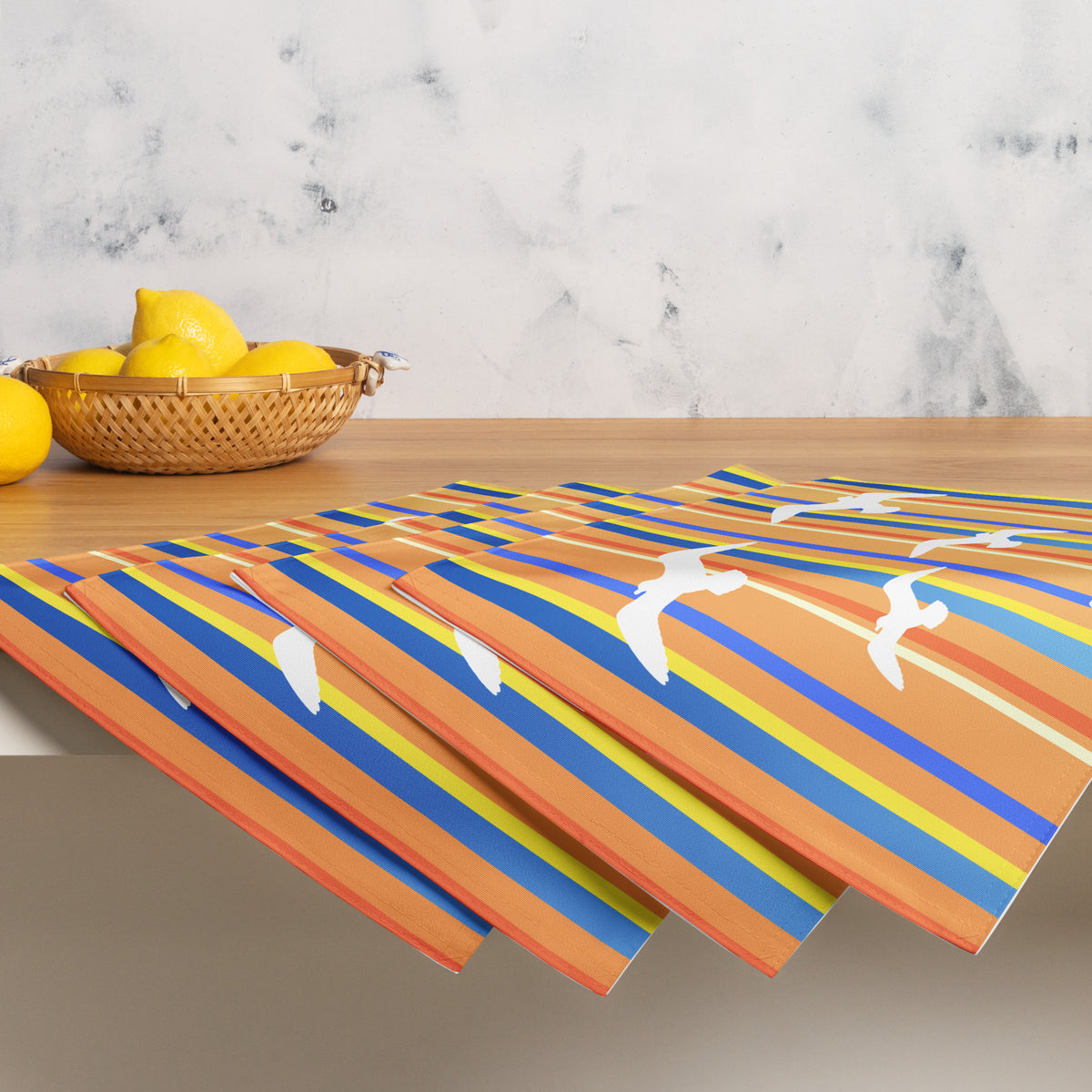 Product mockup of a Petals & Paws Drift Placemat, featuring seagull design with orange striped pattern. This image shows the full set of four with a basket of lemons.