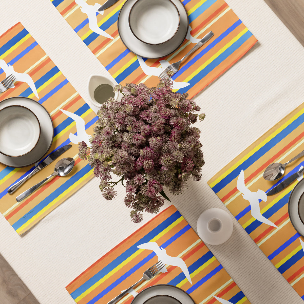 Product mockup of a Petals & Paws Drift Placemat, featuring seagull design with orange striped pattern. This image shows an overhead view of a dining table set for four with plates, bowls, utensils, and a vase holding cut flowers.