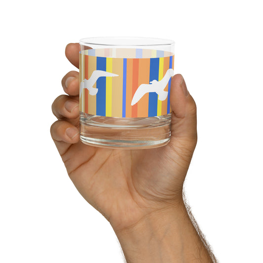 A Petals & Paws Drift Rocks Glass held up in a man's hand against a white background. This glass features a seagull design with orange striped pattern.