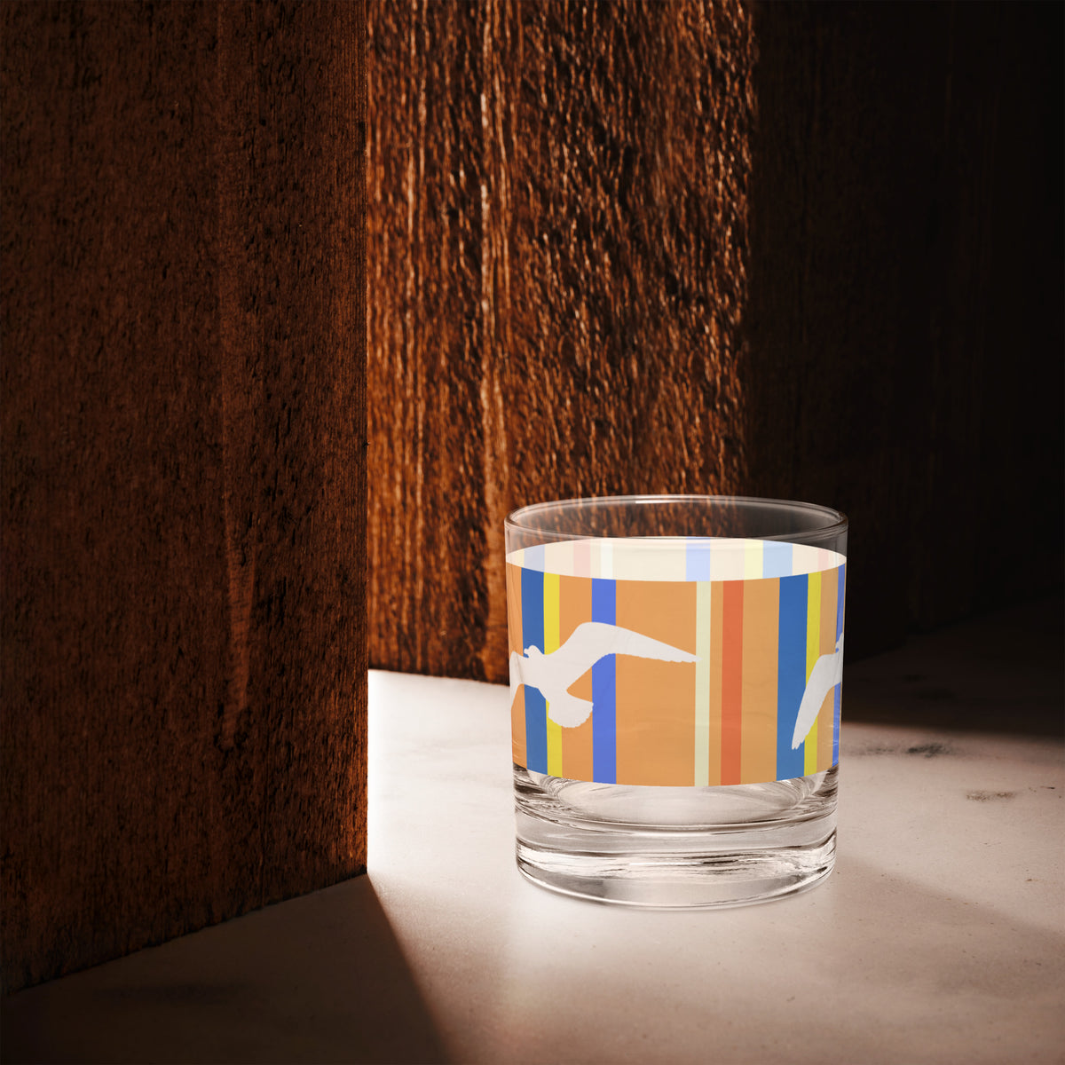 A Petals & Paws Drift Rocks Glass is sitting in the space of light between two cherry-wood-colored walls. This glass features a seagull design with orange striped pattern.