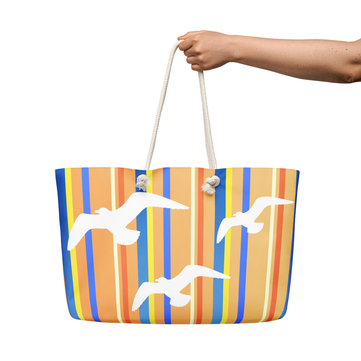 A Petals & Paws Drift Weekender Bag is held out against a white background. The female hand holds the bag by its cream-colored cotton rope handles. This all-over print bag features seagull design with orange striped pattern.