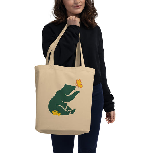Petals & Paws Eco Tote Bag being carried by a woman wearing dark clothes. Oyster cotton, featuring front graphic illustration of a forest green grizzly bear curious about an orange and yellow butterfly while sitting in yellow flowers.  
