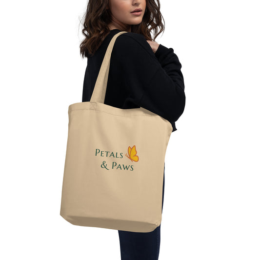 Petals & Paws Eco Tote Bag being carried by a woman wearing dark clothes. Oyster cotton, featuring rear graphic illustration of a forest green text, "Petals & Paws" accented by the brand's orange and yellow butterfly.