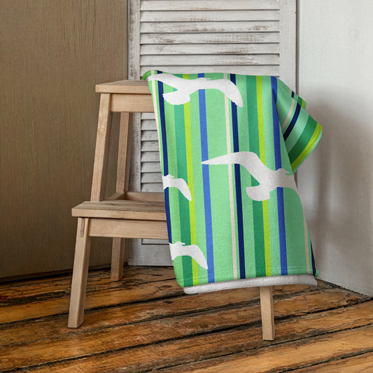 A 30 x 60" Petals & Paws Harbour Beach Towel is draped on a wooden step stool against a neutral wall. This towel features seagull design with green striped pattern.
