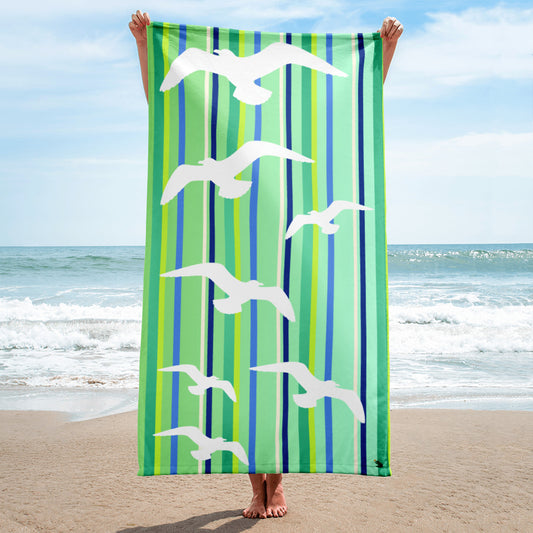 A 30 x 60" Petals & Paws Harbour Beach Towel is held up vertically in front of a woman on a sandy beach. A large body of water is in the background. This towel features seagull design with green striped pattern.