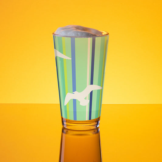In-action mockup of Petals & Paws Harbour Pint Glass, featuring seagull design with green striped pattern. The glass, surrounded by orange glow, is holding beer with frothy foam.