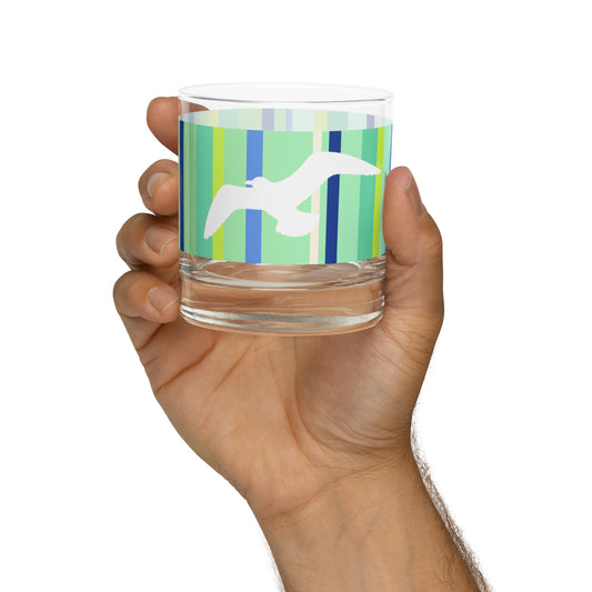 A Petals & Paws Harbour Rocks Glass held up in a man's hand against a white background. This glass features a seagull design with green striped pattern.