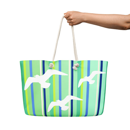 A Petals & Paws Harbour Weekender Bag is held out against a white background. The female hand holds the bag by its cream-colored cotton rope handles. This all-over print bag features seagull design with green striped pattern.