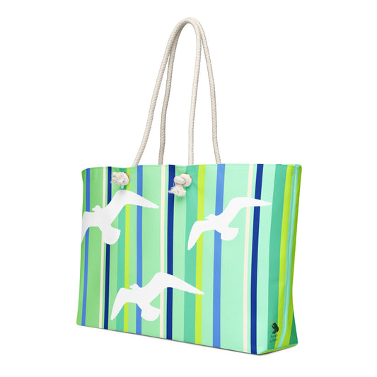 A diagonal view of Petals & Paws Harbour Weekender Bag showing the pattern as it wraps around the ends of the bag, with the Petals & Paws logo near the bottom. This all-over print bag features seagull design with green striped pattern and cream-colored cotton rope handles.