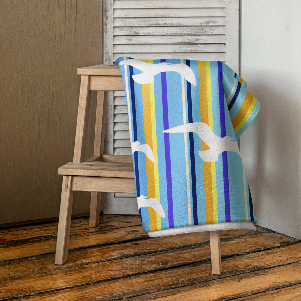 A 30 x 60" Petals & Paws Shoreline Beach Towel is draped on a wooden step stool against a neutral wall. This towel features seagull design with blue striped pattern.