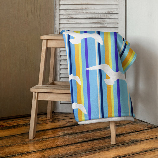 A 30 x 60" Petals & Paws Shoreline Beach Towel is draped on a wooden step stool against a neutral wall. This towel features seagull design with blue striped pattern.