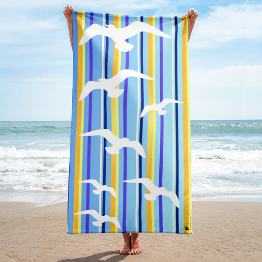 A 30 x 60" Petals & Paws Shoreline Beach Towel is held up vertically in front of a woman on a sandy beach. A large body of water is in the background. This towel features seagull design with blue striped pattern.