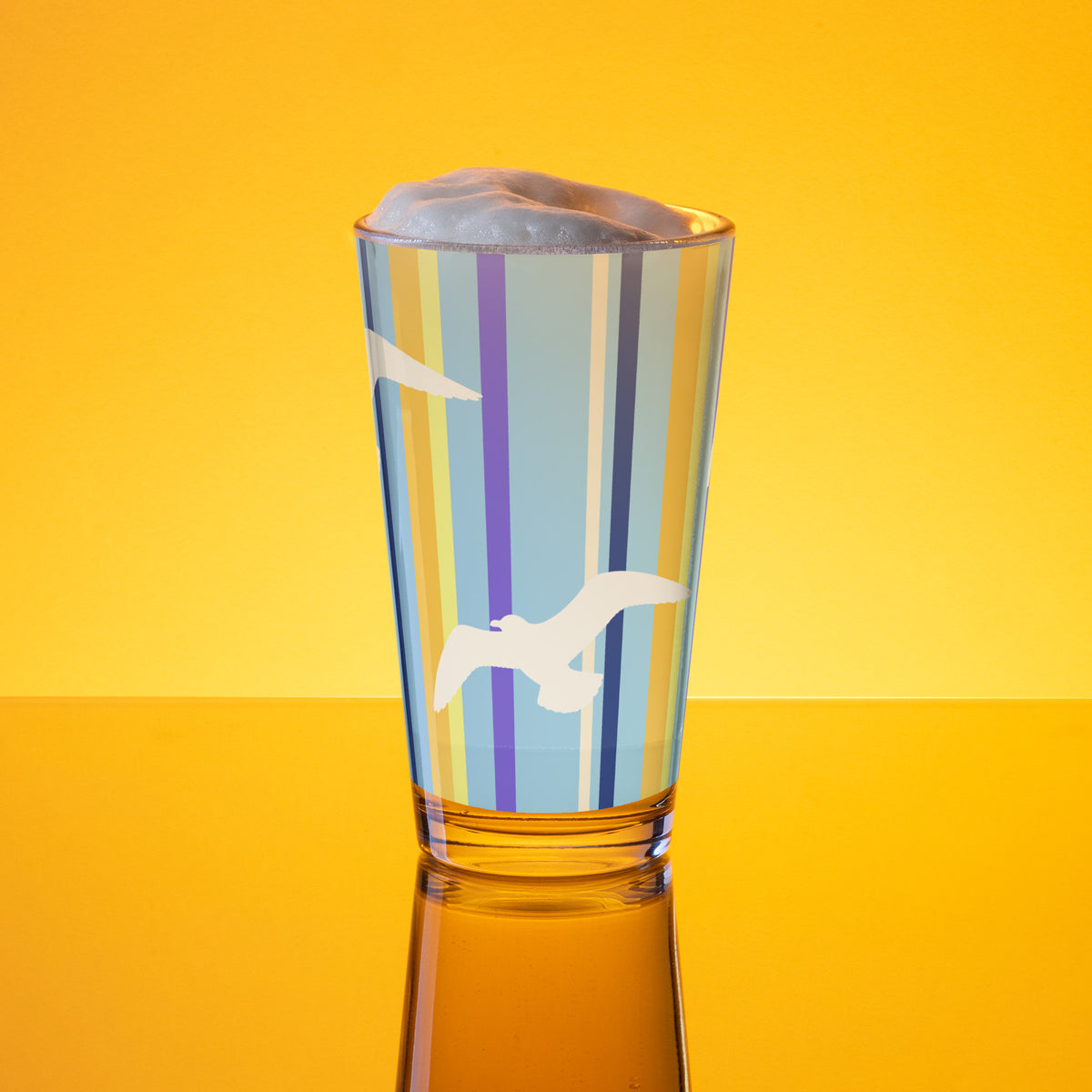 In-action mockup of Petals & Paws Shoreline Pint Glass, featuring seagull design with blue striped pattern. The glass, surrounded by orange glow, is holding beer with frothy foam.
