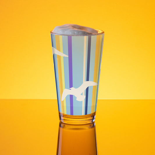 In-action mockup of Petals & Paws Shoreline Pint Glass, featuring seagull design with blue striped pattern. The glass, surrounded by orange glow, is holding beer with frothy foam.