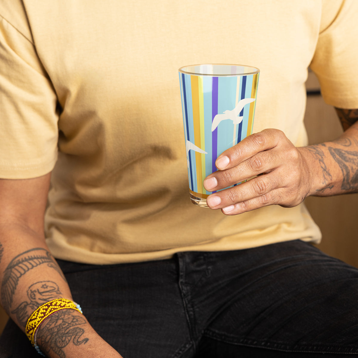 A man holding the Petals & Paws Shoreline Pint Glass, featuring seagull design with blue striped pattern. Chest and tattooed arms are within the picture.