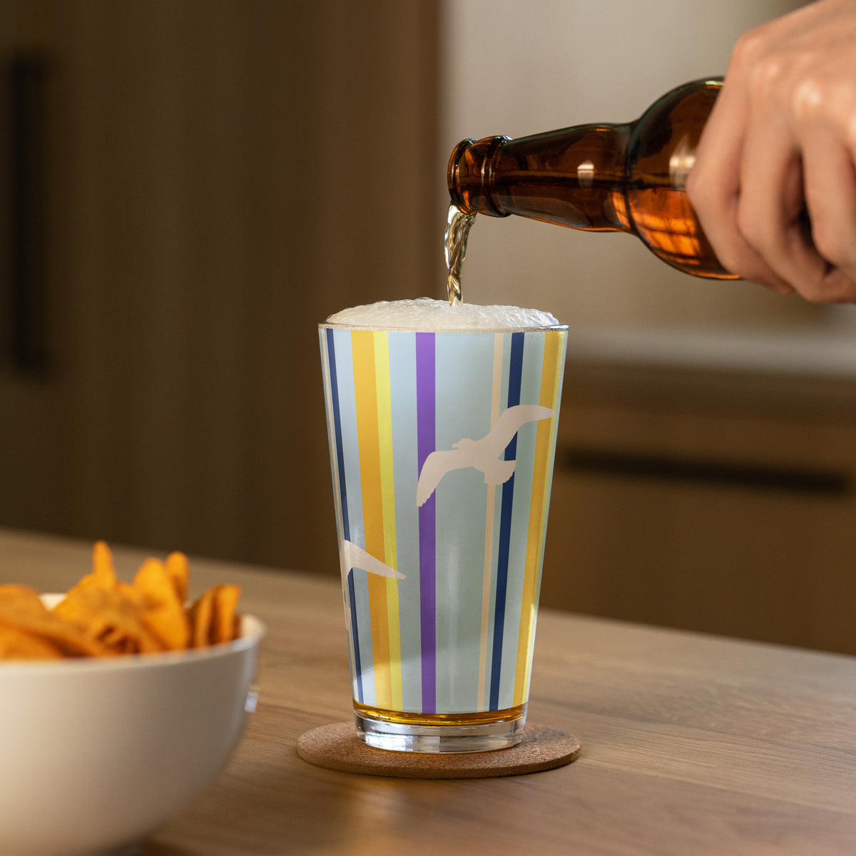 In-action mockup of Petals & Paws Shoreline Pint Glass, featuring seagull design with blue striped pattern. A hand is pouring beer into the pint glass and a bowl of nachos sits nearby.