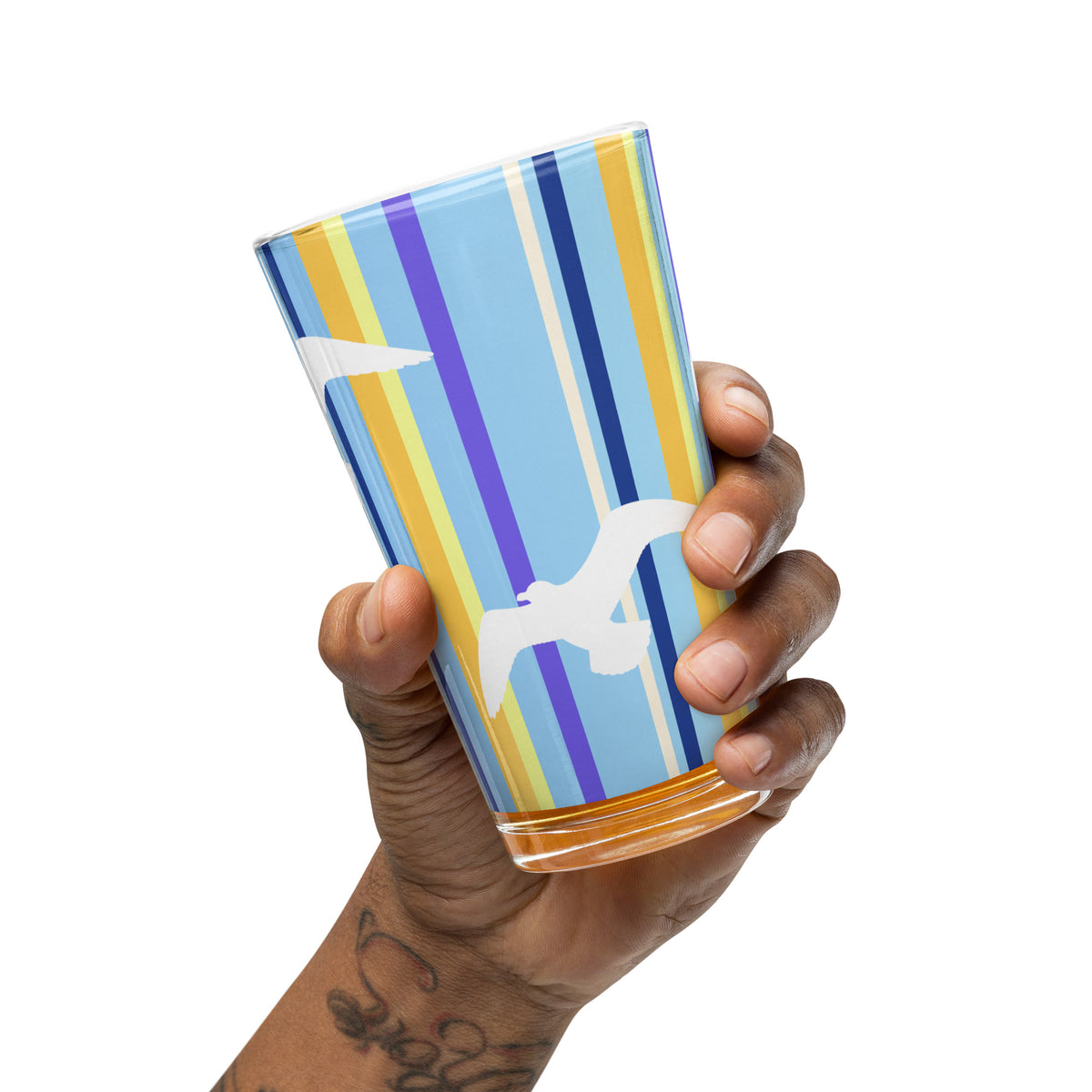 A hand extended from a tattooed arm tips the Petals & Paws Shoreline Pint Glass slightly.