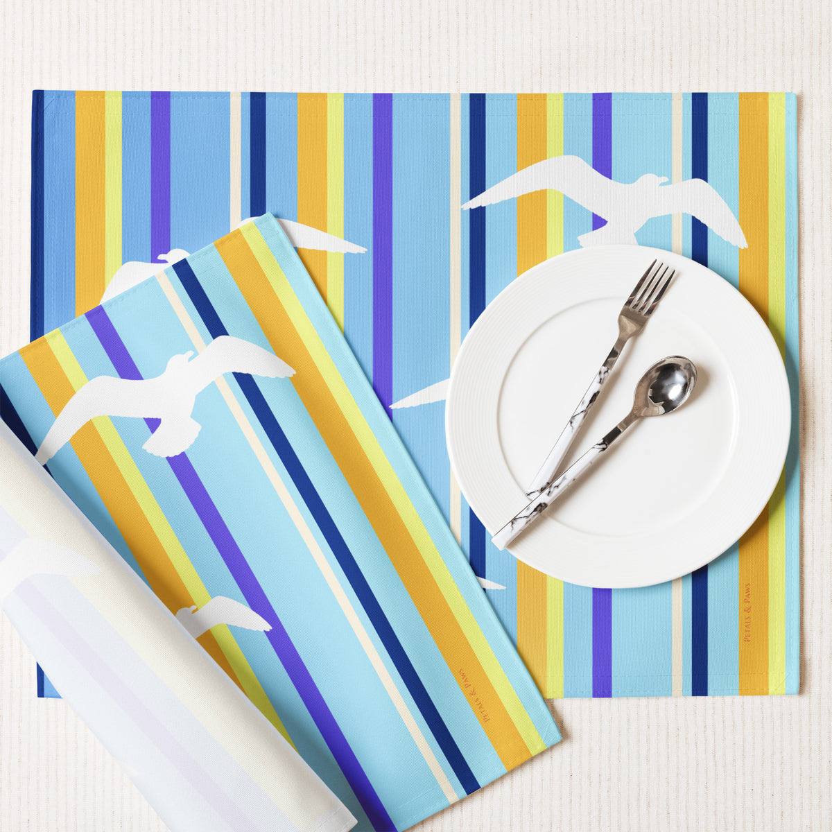 Product mockup of a Petals & Paws Shoreline Placemat, featuring seagull design with blue striped pattern. This image shows the white back and hemmed edges.
