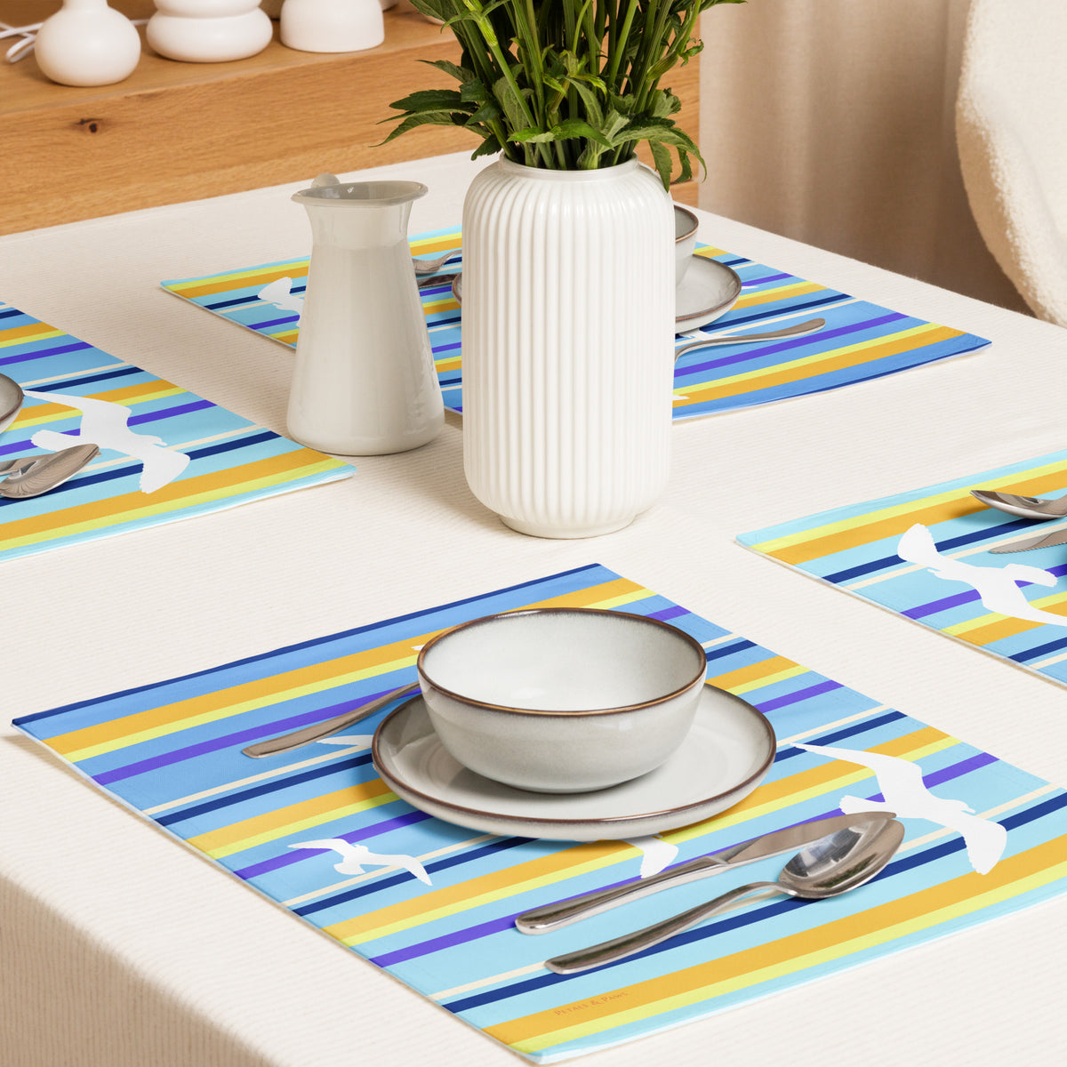 Product mockup of a Petals & Paws Shoreline Placemat, featuring seagull design with blue striped pattern. This image shows cross-view of a dining table set for four with plates, bowls, utensils, and a vase holding cut flowers.
