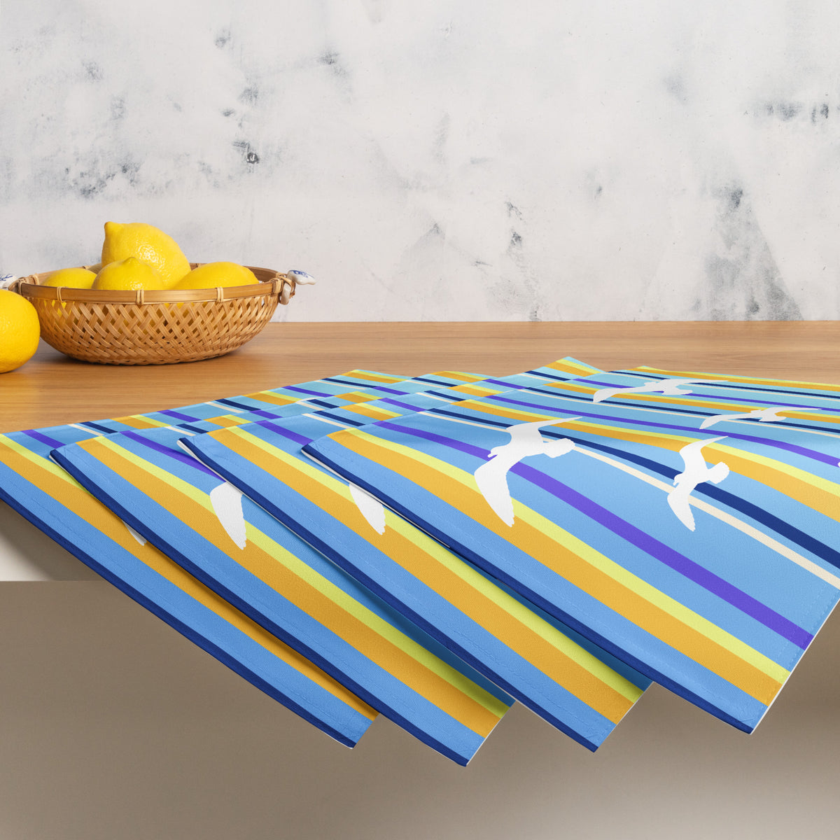 Product mockup of a Petals & Paws Shoreline Placemat, featuring seagull design with blue striped pattern. This image shows the full set of four with a basket of lemons.