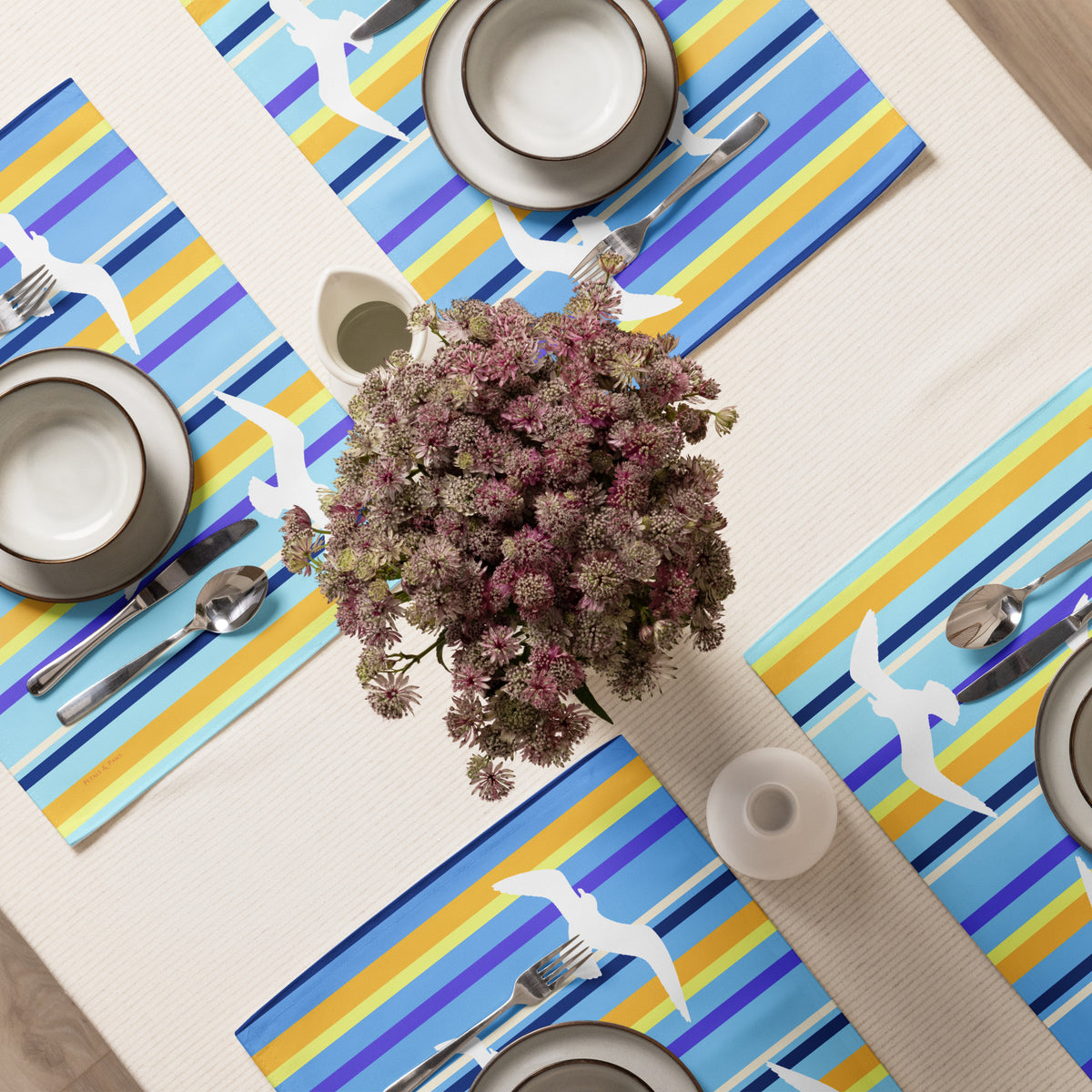 Product mockup of a Petals & Paws Shoreline Placemat, featuring seagull design with blue striped pattern. This image shows an overhead view of a dining table set for four with plates, bowls, utensils, and a vase holding cut flowers.