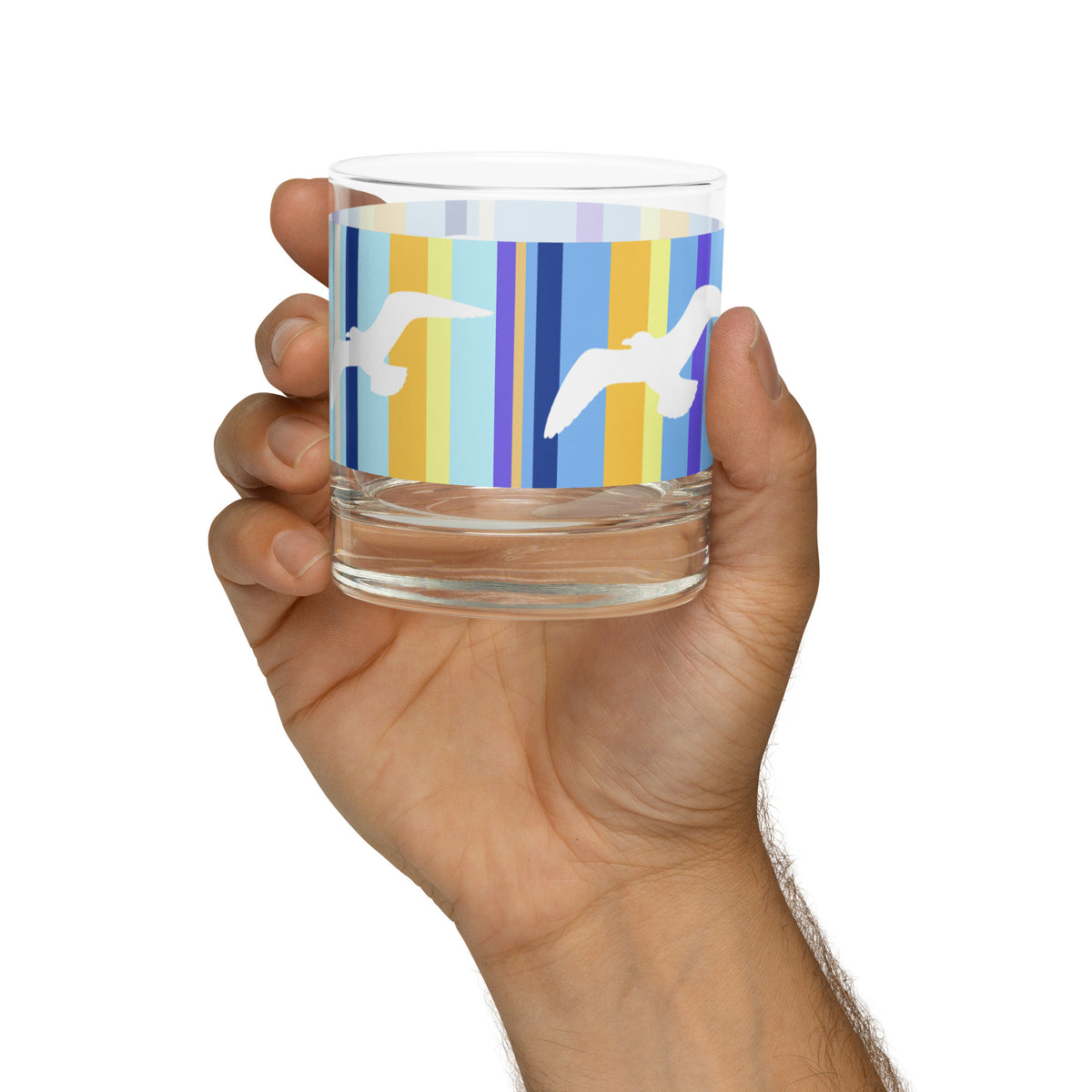 A Petals & Paws Shoreline Rocks Glass held up in a man's hand against a white background. This glass features a seagull design with blue striped pattern.