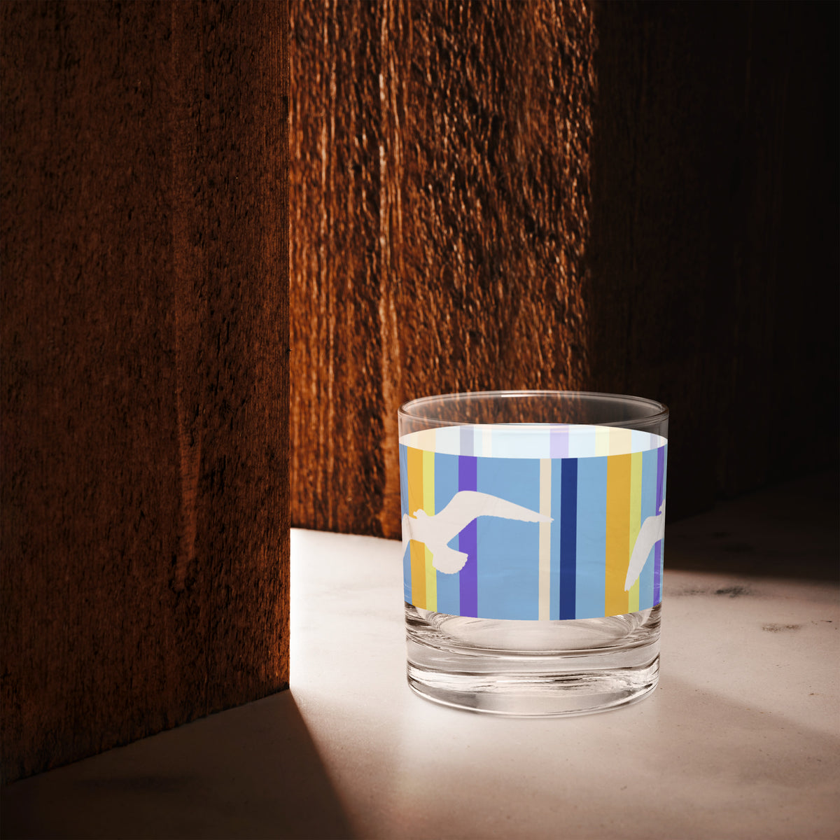 A Petals & Paws Shoreline Rocks Glass is sitting in the space of light between two cherry-wood-colored walls. This glass features a seagull design with blue striped pattern.