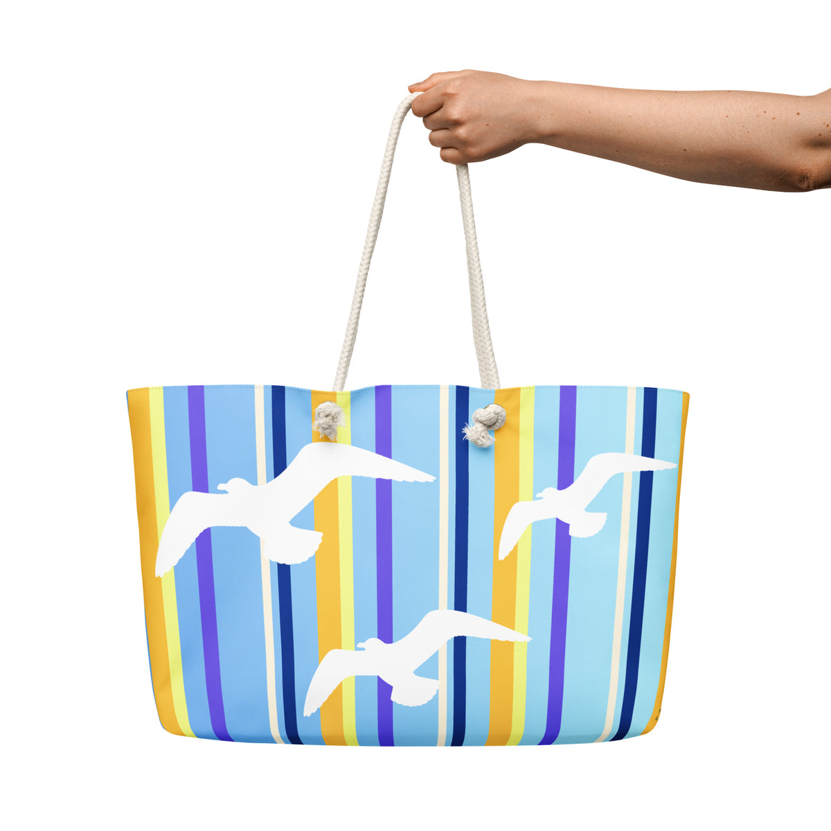 A Petals & Paws Shoreline Weekender Bag is held out against a white background. The female hand holds the bag by its cream-colored cotton rope handles. This all-over print bag features seagull design with blue striped pattern.
