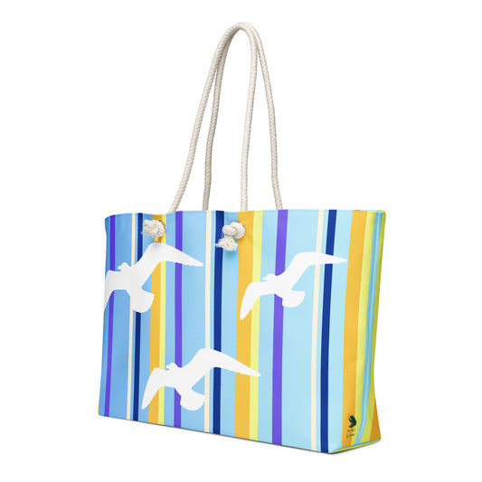 A diagonal view of Petals & Paws Shoreline Weekender Bag showing the pattern as it wraps around the ends of the bag, with the Petals & Paws logo near the bottom. This all-over print bag features seagull design with blue striped pattern and cream-colored cotton rope handles.