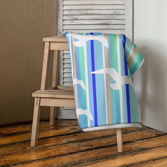 A 30 x 60" Petals & Paws Tide Beach Towel is draped on a wooden step stool against a neutral wall. This towel features seagull design with blue striped pattern.