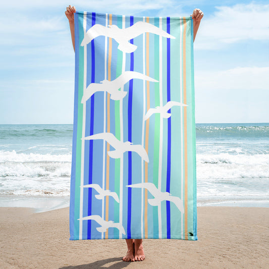 A 30 x 60" Petals & Paws Tide Beach Towel is held up vertically in front of a woman on a sandy beach. A large body of water is in the background. This towel features seagull design with blue striped pattern.