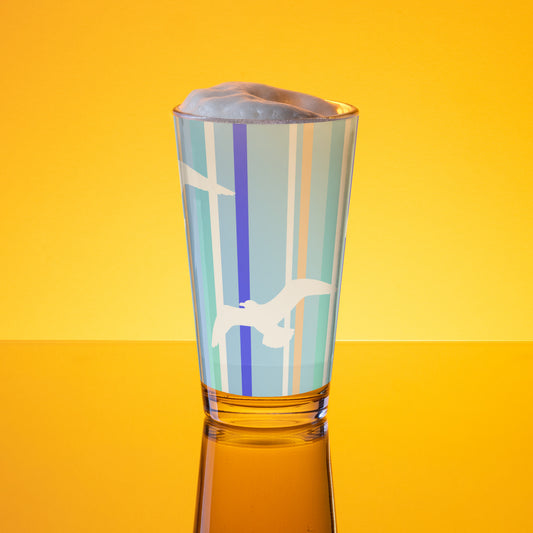 In-action mockup of Petals & Paws Tide Pint Glass, featuring seagull design with blue striped pattern. The glass, surrounded by orange glow, is holding beer with frothy foam.