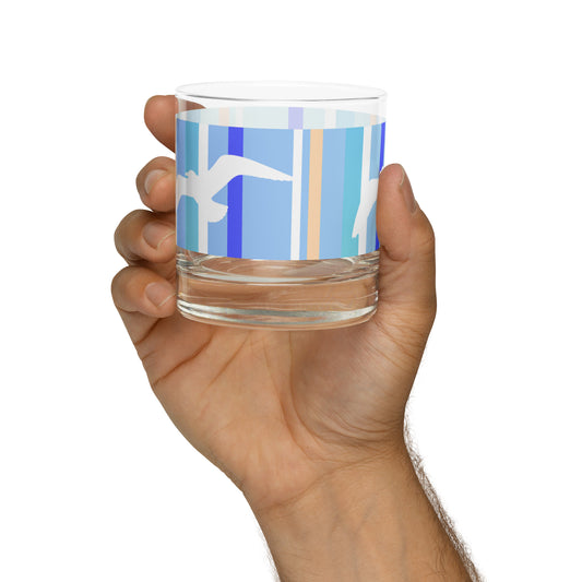 A Petals & Paws Tide Rocks Glass held up in a man's hand against a white background. This glass features a seagull design with blue striped pattern.
