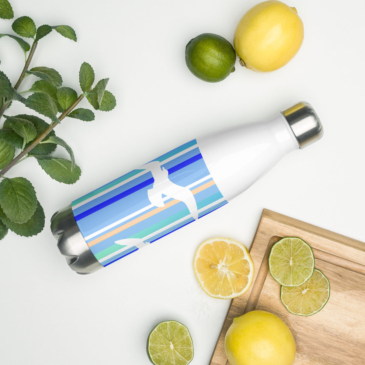 A 17 oz Petals & Paws Tide Water Bottle laying on its side amidst citrus fruit and an herb plant. This bottle features seagull design with blue striped pattern.