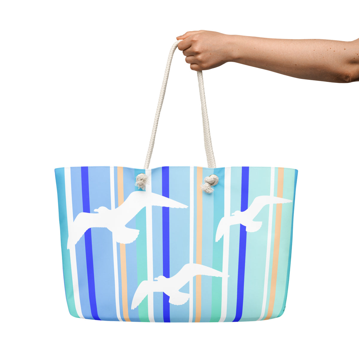 A Petals & Paws Tide Weekender Bag is held out against a white background. The female hand holds the bag by its cream-colored cotton rope handles. This all-over print bag features seagull design with blue striped pattern.