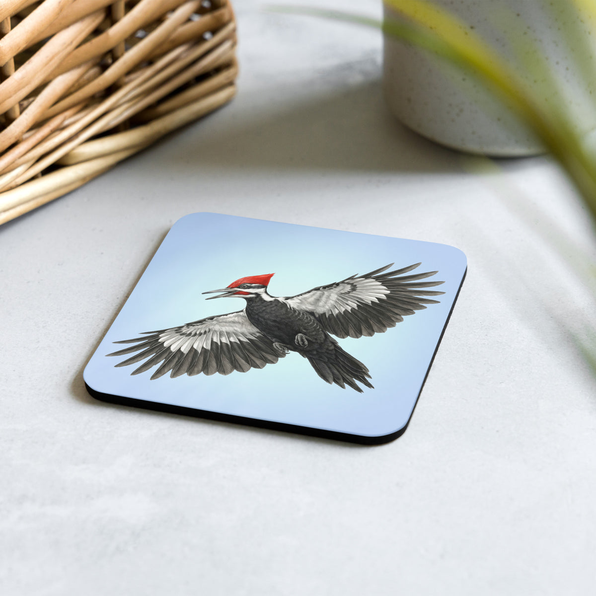Product mockup of Pileated Woodpecker Coaster set on a desk with a woven basket and clay mug. Design is a woodpecker is flying against a clear blue sky.