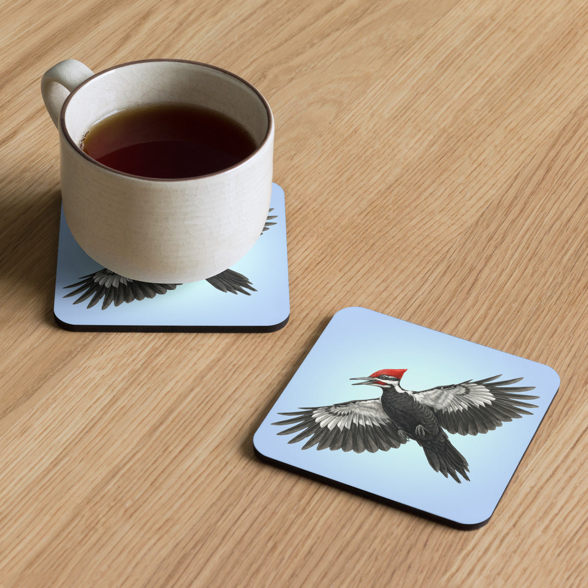 Product mockup of two Pileated Woodpecker Coasters, one with a mug of tea sitting on it, and the other set beside, both on a faux-wood surface. Design is a woodpecker is flying against a clear blue sky.