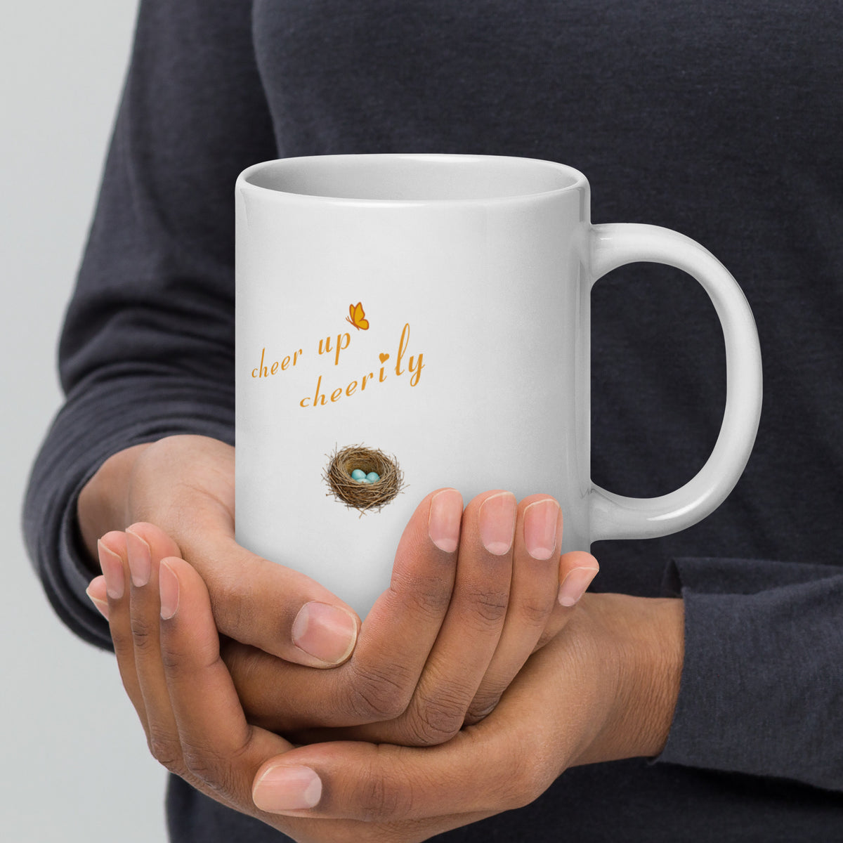 A man's hands hold a 20 oz Robin Discovery Ceramic Mug.  Back design of nest with robin-blue eggs, "cheer up cheerily," set against a dark sweater.