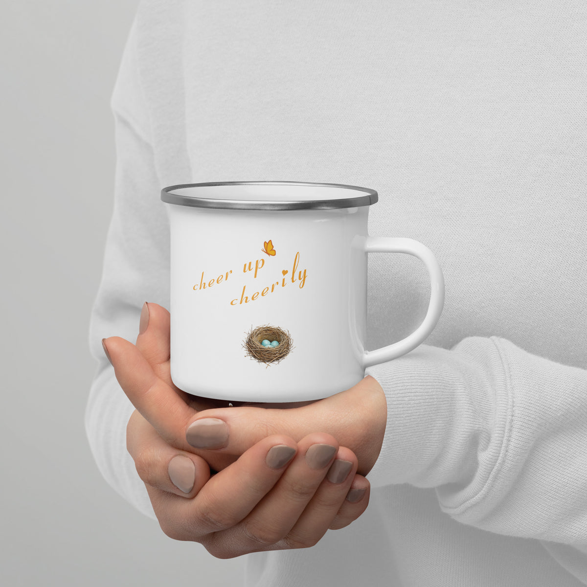 In-action shot of Robin Discovery Enamel Mug held in a woman's hands against a white sweater.  Back design, a nest of robin-blue eggs and, "cheer up cheerily."