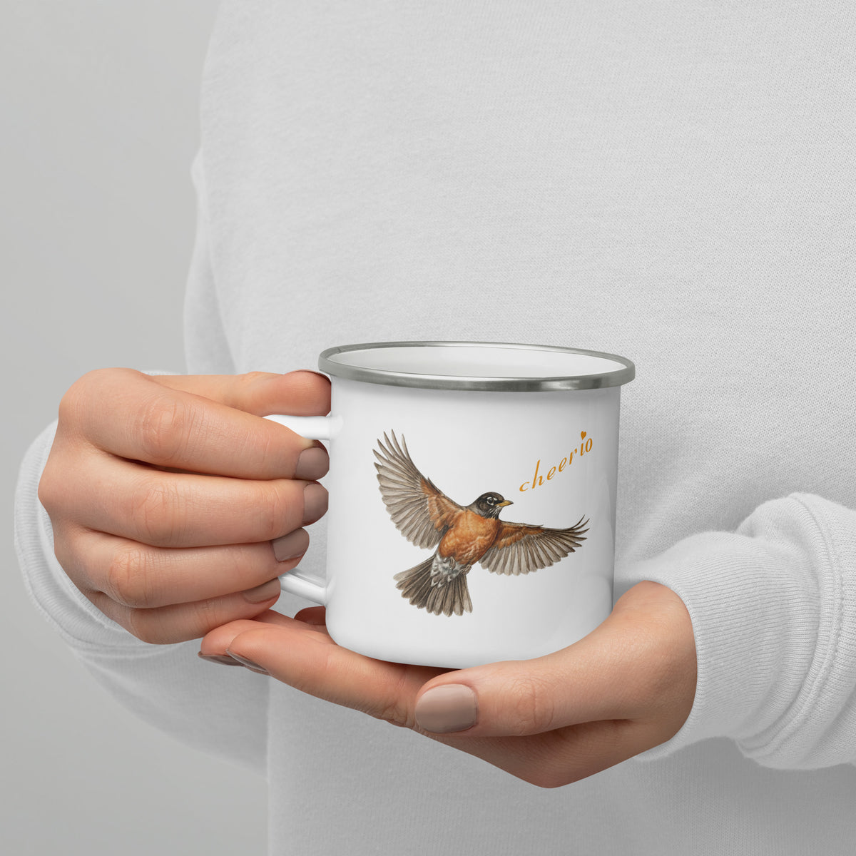 In-action shot of Robin Discovery Enamel Mug held in a woman's hands against a white sweater.  Front design, a realistic flying robin and, "cheerio."