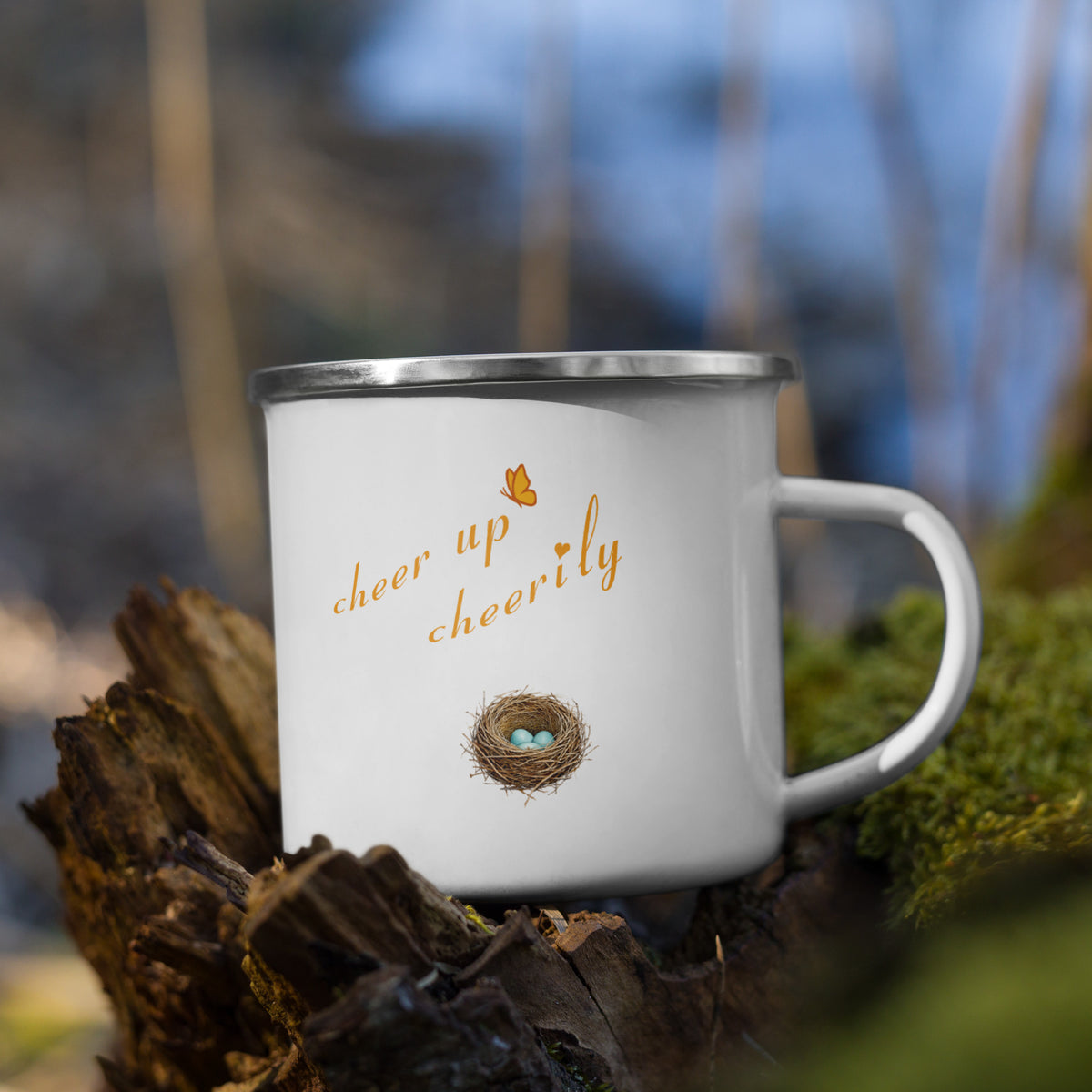 Product mockup of Robin Discovery Enamel Mug sitting in a moss-covered stump and revealing back design, a nest of robin-blue eggs and, "cheer up cheerily."