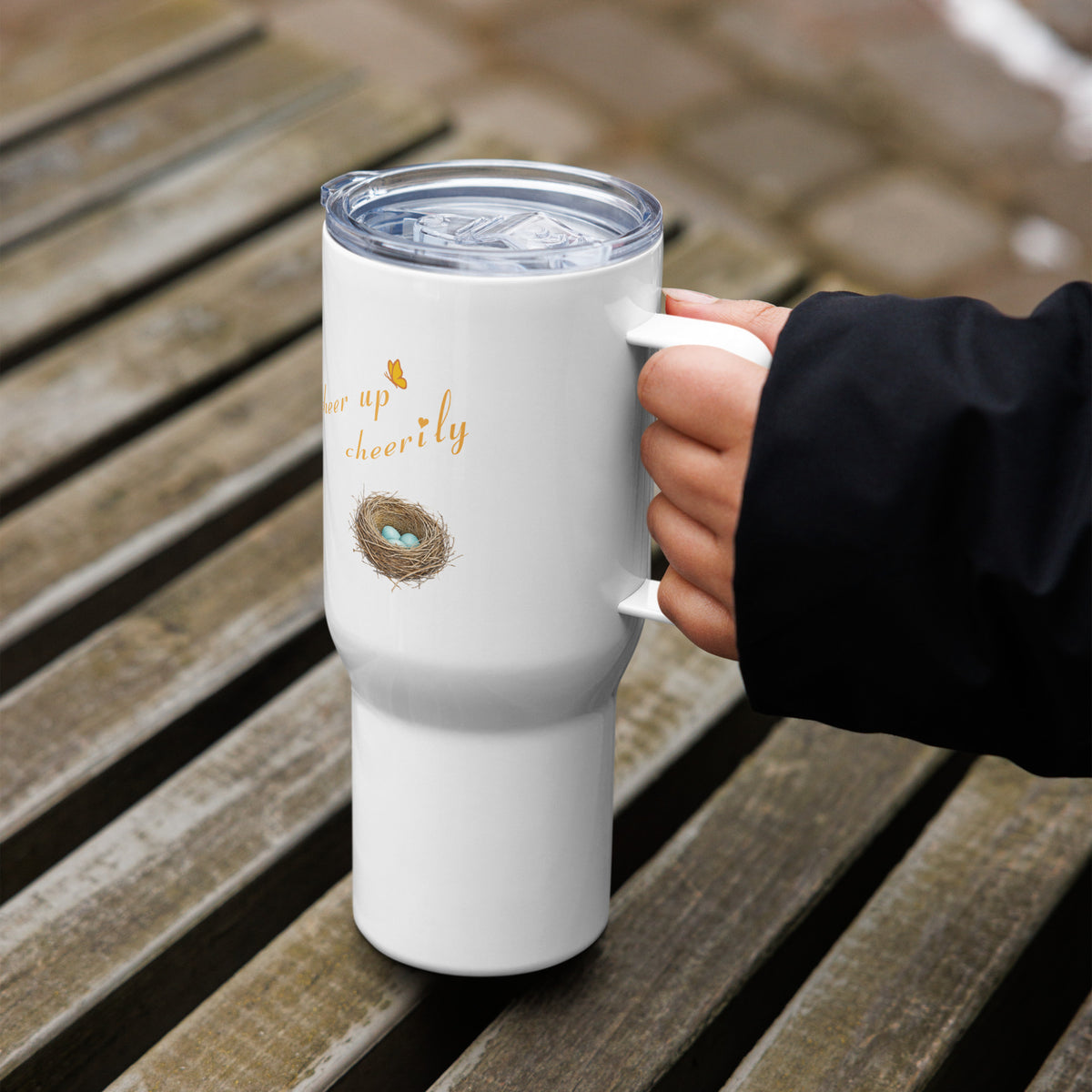 In-action shot, 25 oz Robin Discovery Travel Mug held in a hand with a wooden backdrop. Lid and handle, and back-facing design is a nest with 3 robin-blue eggs and, "cheer up cheerily" against a white background.
