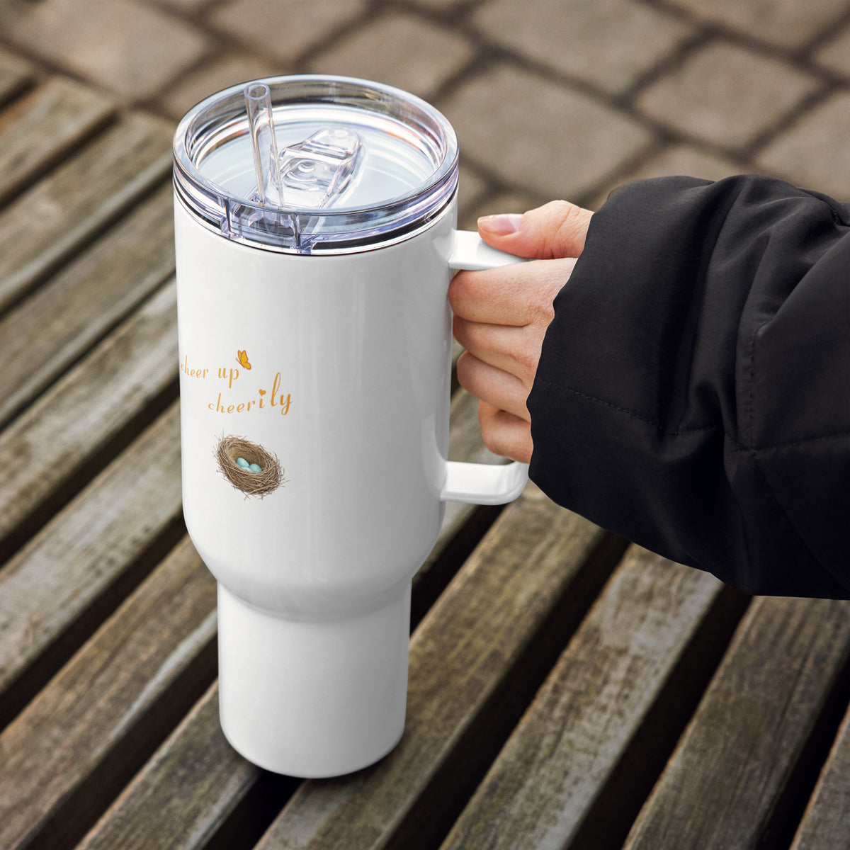 In-action shot, 40 oz Robin Discovery Travel Mug held in a hand with a wooden backdrop. Lid, handle, and straw, and back-facing design is a nest with 3 robin-blue eggs and, "cheer up cheerily" against a white background.