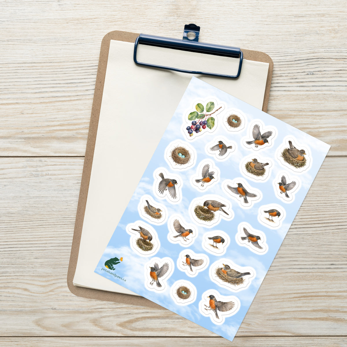 A sheet of kiss-cut stickers featuring illustrated robins nesting and in flight with a fair blue sky background, placed on top of an open clipboard with a blank sheet on a light wood tabletop.