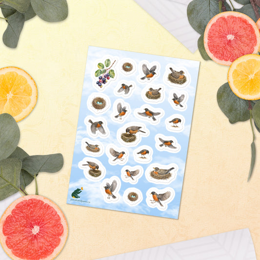 A sheet of kiss-cut stickers featuring illustrated American robins nesting and in flight, with a background of fair blue sky, placed next to citrus slices on a warm-colored background.
